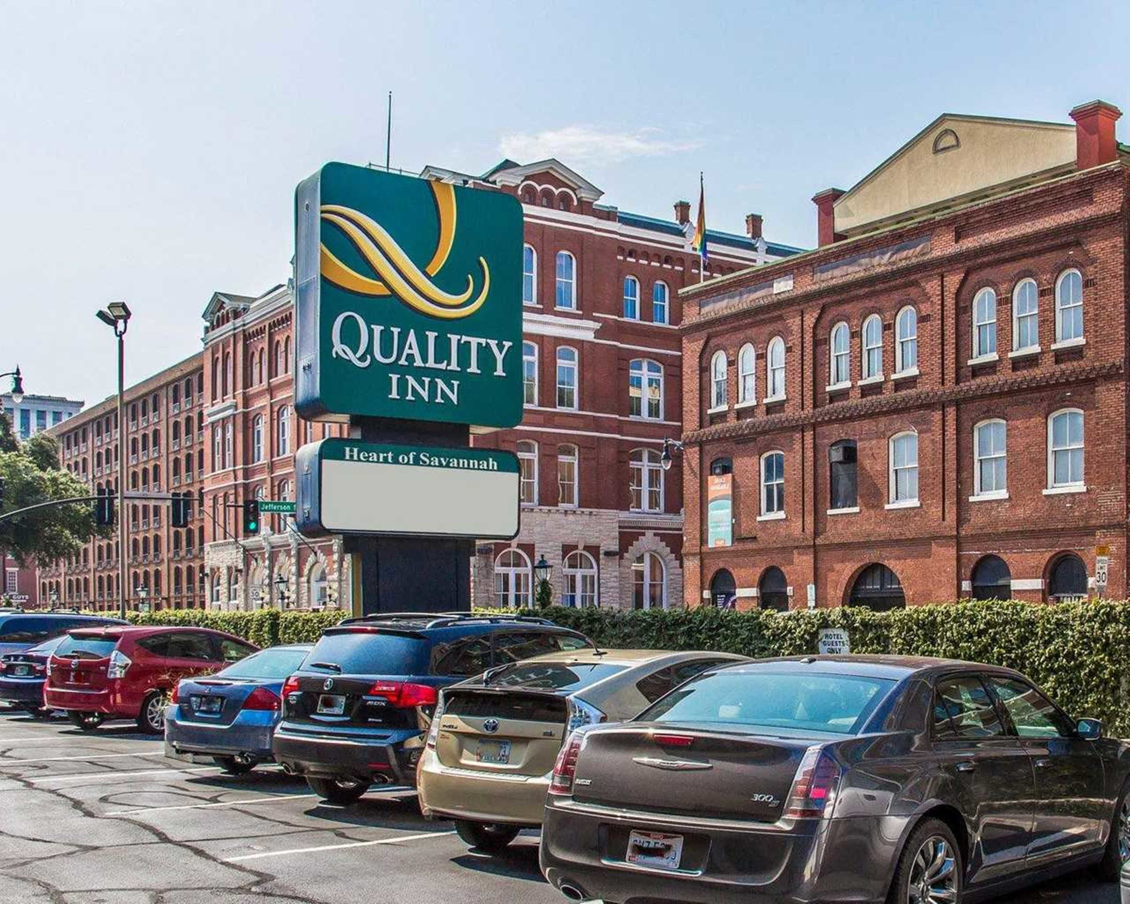 A Quality Inn sign with "Heart of Savannah" text, surrounded by parked cars and historic brick buildings.