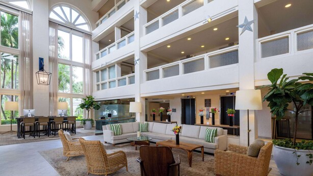 A spacious lobby with a high ceiling, multiple seating areas, and large windows offering views of palm trees.