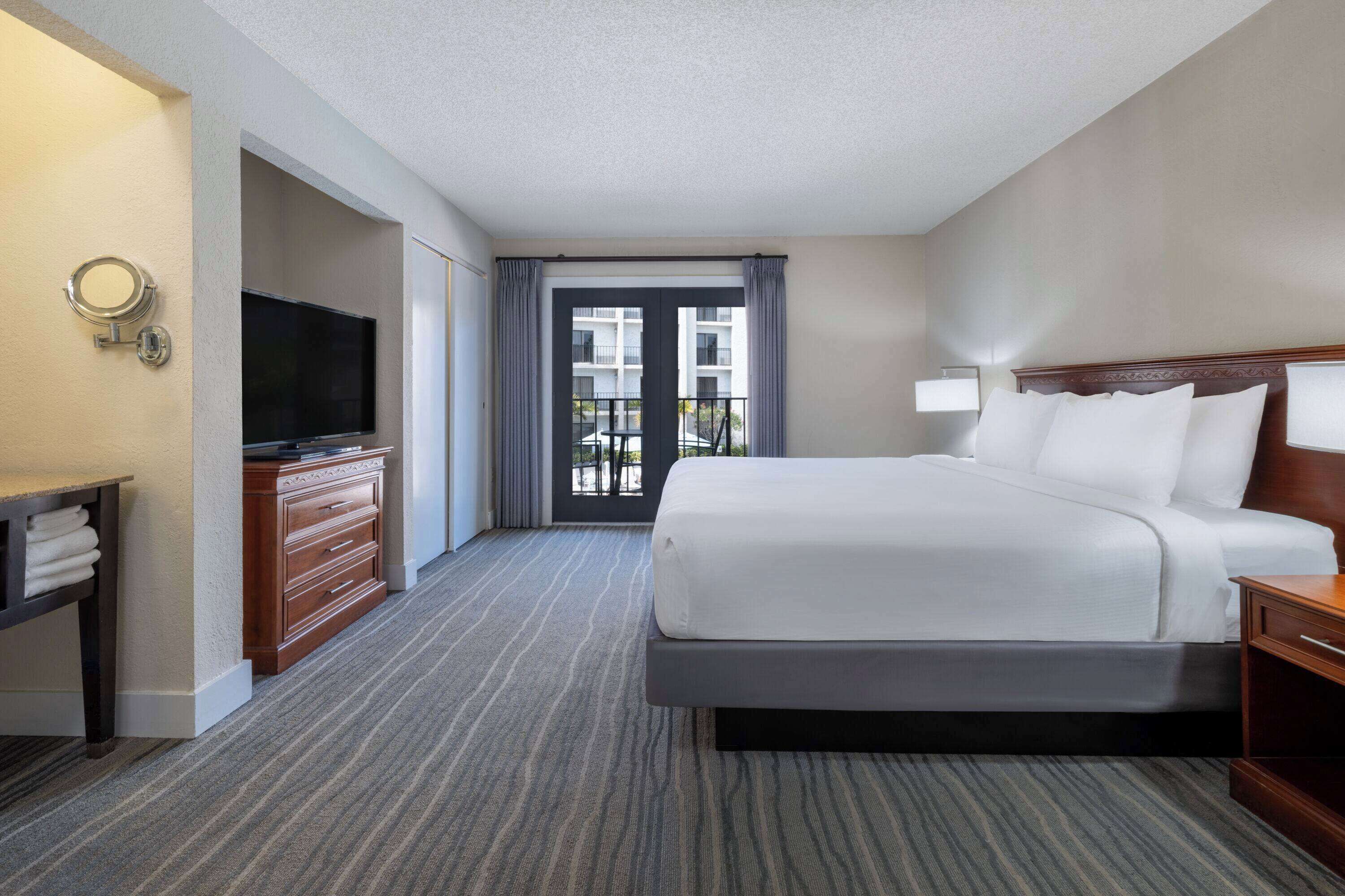 A hotel room with a large bed, a television, a dresser, and a balcony with a view of the building.