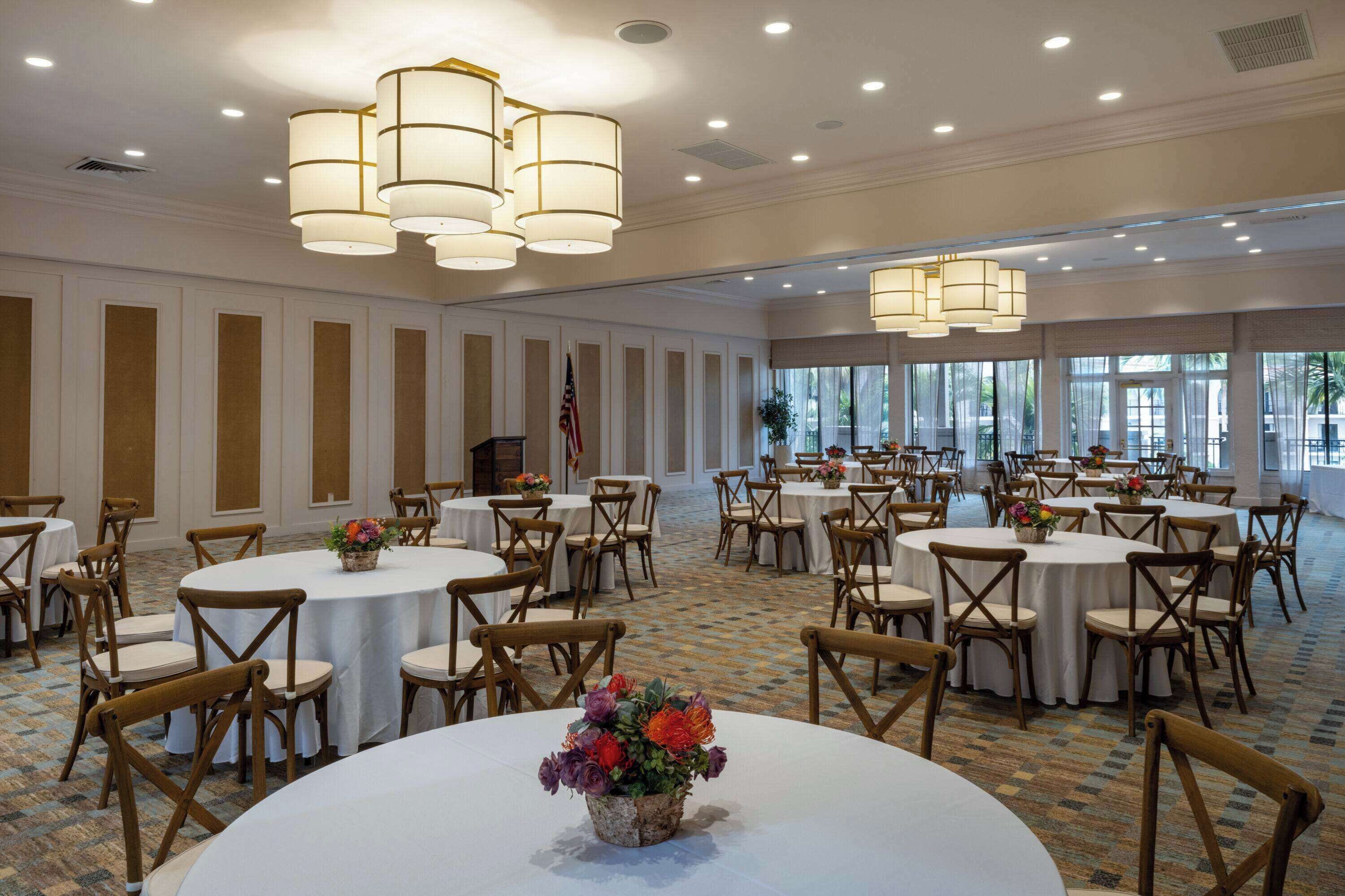 A large banquet hall with round tables and chairs, chandeliers, and a flag.