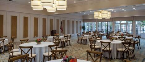 A large banquet hall with round tables and chairs, chandeliers, and a flag.