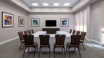 Una sala de conferencias con una mesa larga, sillas marrones, obras de arte enmarcadas en las paredes, un televisor de pantalla plana y ventanas grandes.