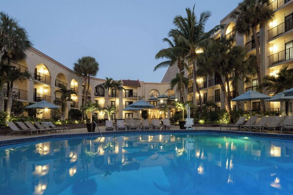 A hotel courtyard with a swimming pool, sun loungers, palm trees, and a well-lit building facade. - Wyndham Boca Raton Hotel (Boca Raton)