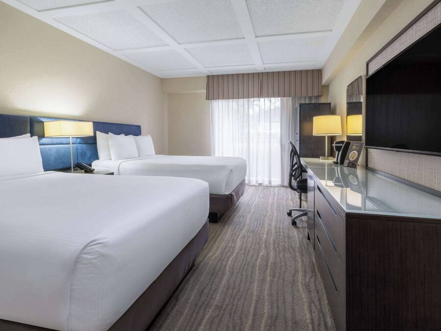 A hotel room with two beds, a desk with a flat-screen TV, and a large window with curtains.