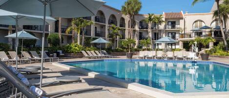 A pool area with lounge chairs and umbrellas, surrounded by a multi-story building with balconies and palm trees.