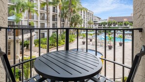 A balcony with a round table, overlooking a pool area with palm trees and a building with balconies.