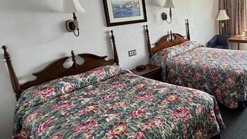 A hotel room with two wooden beds, floral bedding, a framed picture on the wall, and a small table with a chair.