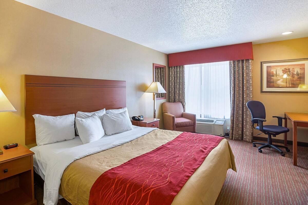 A hotel room with a large bed, a desk, a chair, a nightstand, a lamp, and a window with curtains.