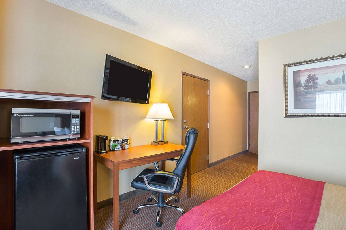 A hotel room with a bed, a desk with a lamp, a microwave, a refrigerator, a TV, and a door.