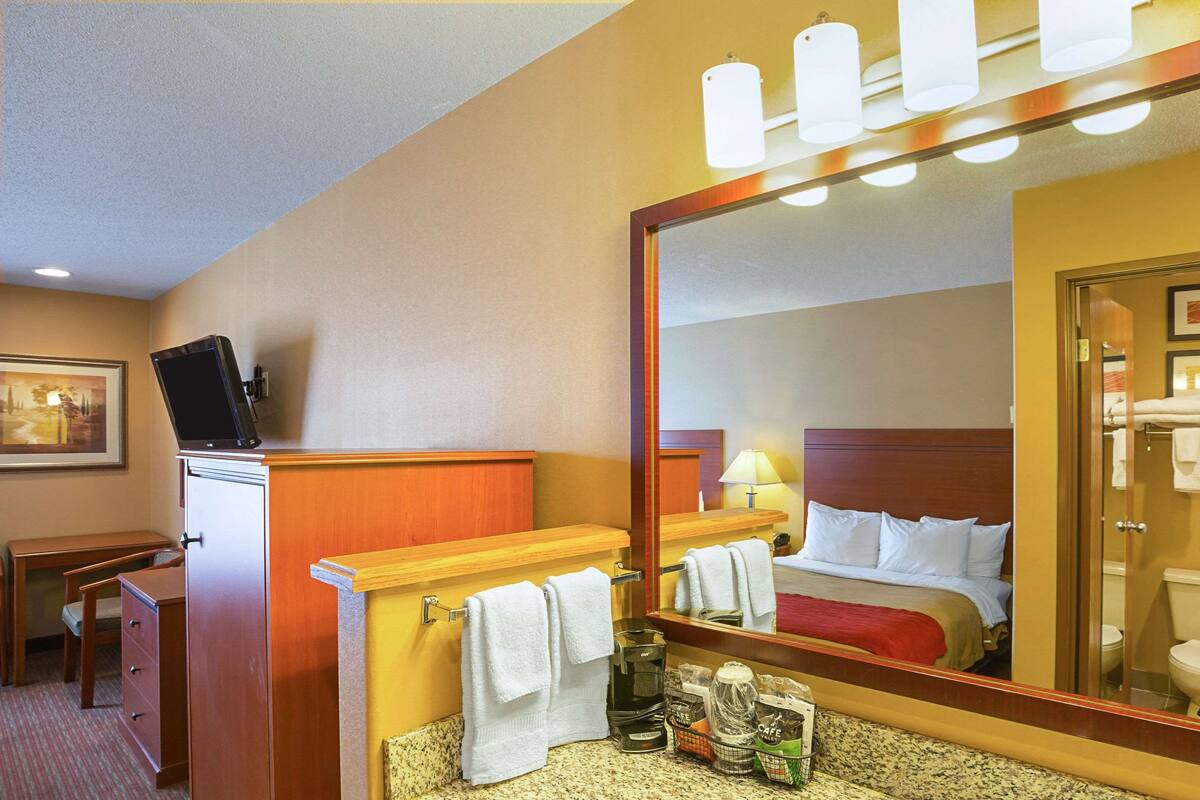 A hotel bathroom with a sink, mirror, and a bed visible in the reflection.