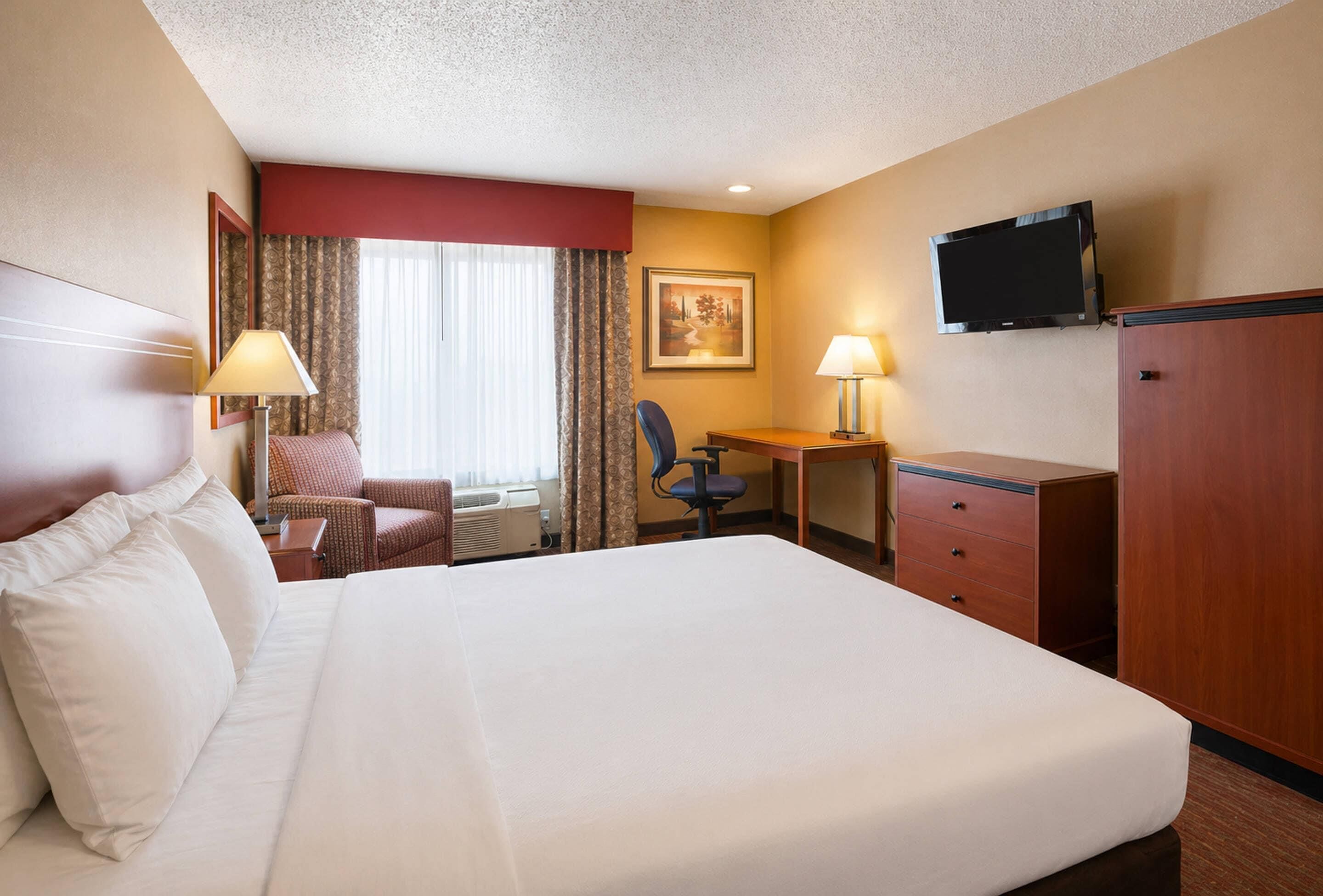 A hotel room with a large bed, a desk, a chair, a television, and a wardrobe.