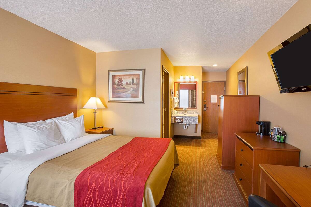 A hotel room with a bed, a desk, a TV, a bathroom, and a painting on the wall.