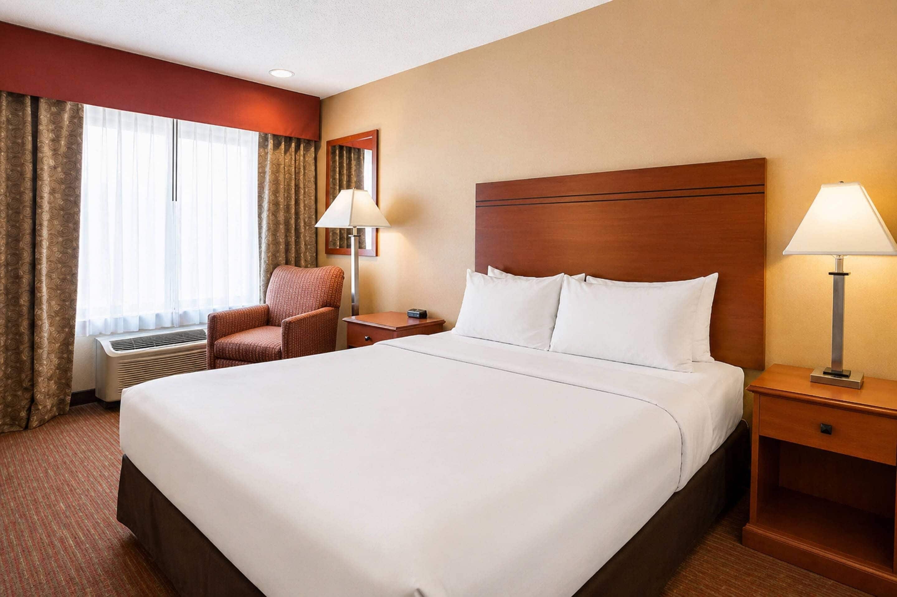 A hotel room with a large bed, a chair, a nightstand, a lamp, and a window with curtains.
