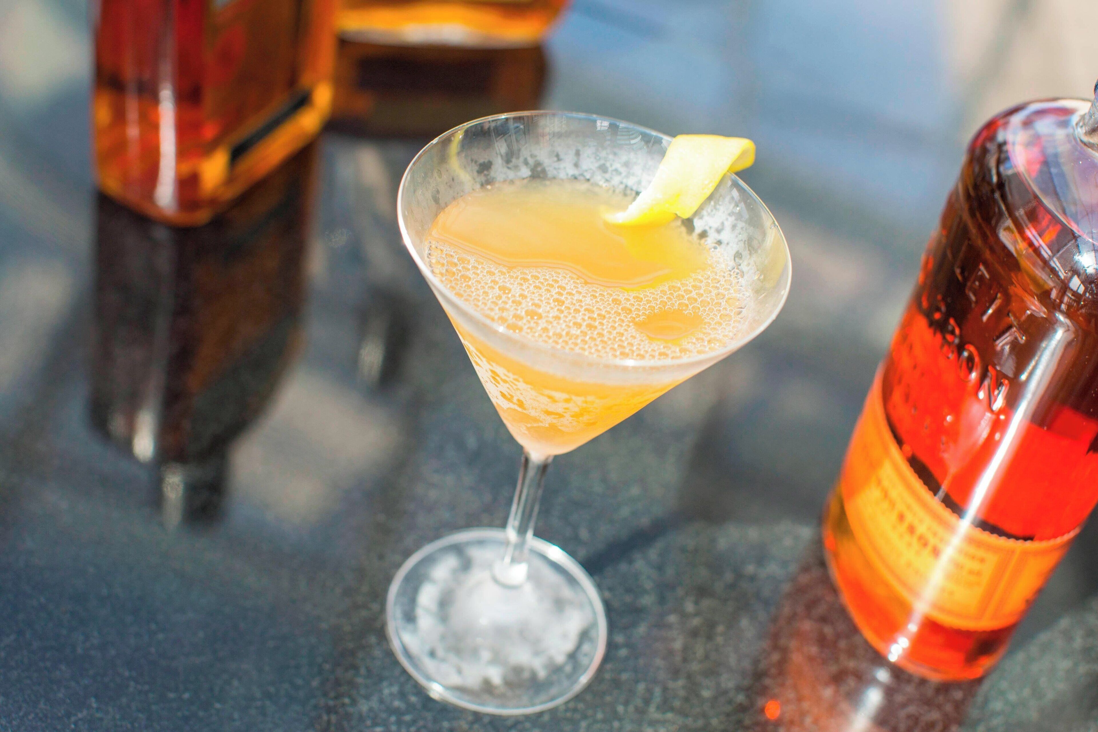 Un cocktail servi dans un verre à martini, garni d’une tranche de citron, une bouteille de jus d’orange et une bouteille contenant un liquide rouge, le tout posé sur une table.
