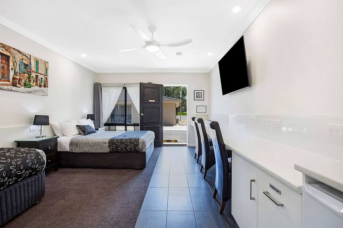 A modern hotel room with a bed, bedside tables, a flat-screen TV, a ceiling fan, and a view of the outside.