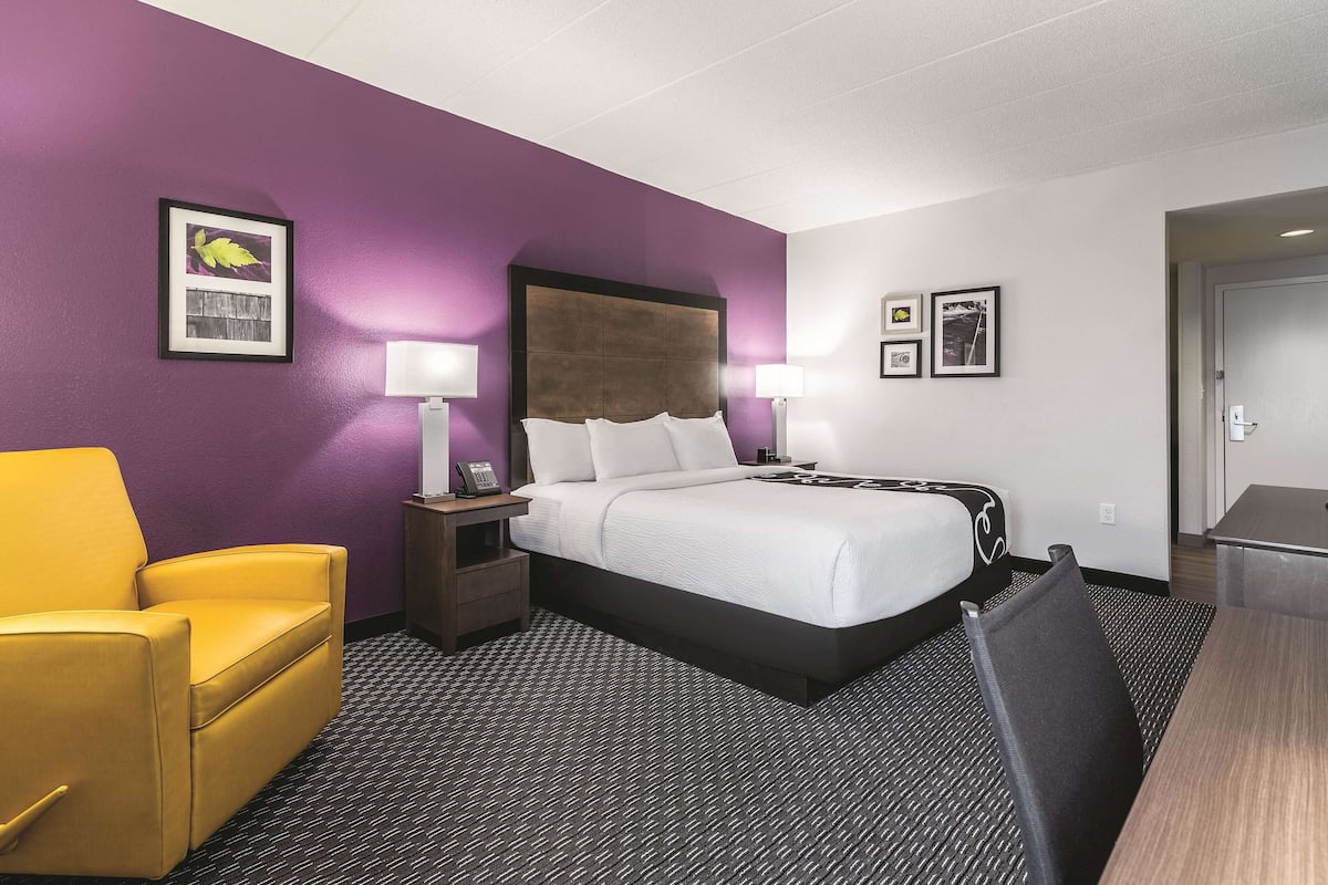 A hotel room with a bed, a desk, a chair, and a nightstand with a lamp.