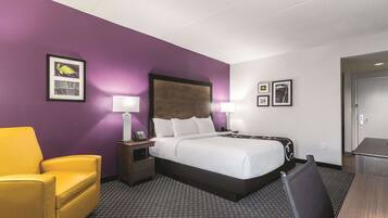 A hotel room with a bed, a desk, a chair, and a nightstand with a lamp.