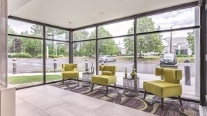 A modern hotel lobby with large windows, yellow chairs, a patterned rug, and a view of the street outside.