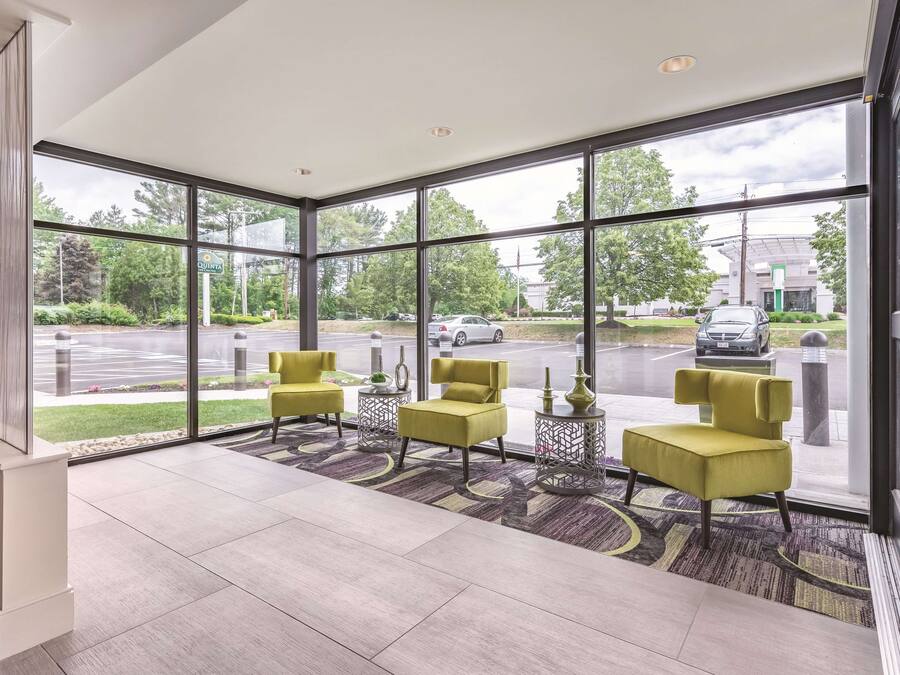 A modern hotel lobby with large windows, yellow chairs, a patterned rug, and a view of the street outside.
