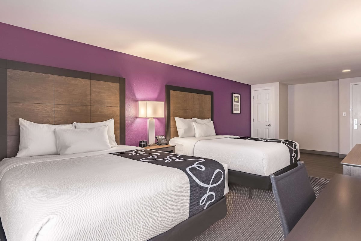 A hotel room with two beds, a nightstand with a lamp, a door, and a small table.