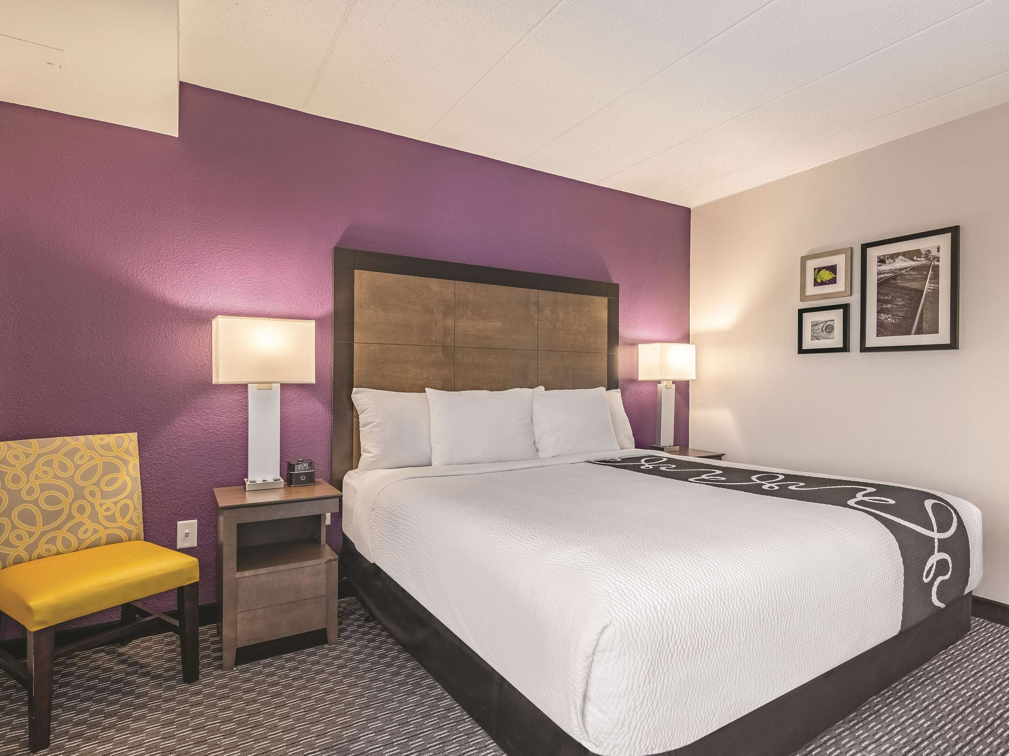 A hotel room with a bed, two bedside lamps, a nightstand, a chair, and a wall with framed pictures.