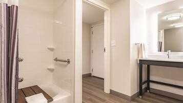 A bathroom with a shower, a wooden bench, a towel, a sink, and a mirror.