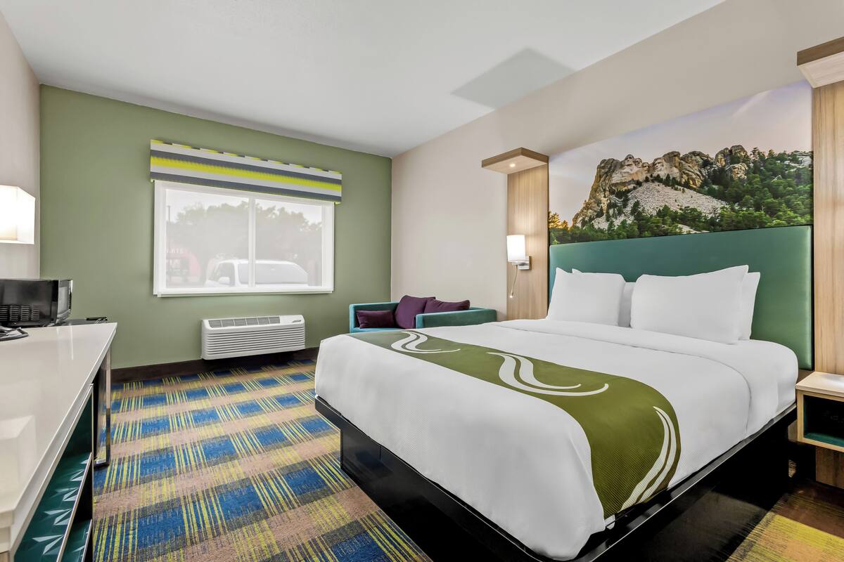 A hotel room with a bed, a desk, a chair, a window with blinds, a wall-mounted air conditioner, and a mural on the wall.