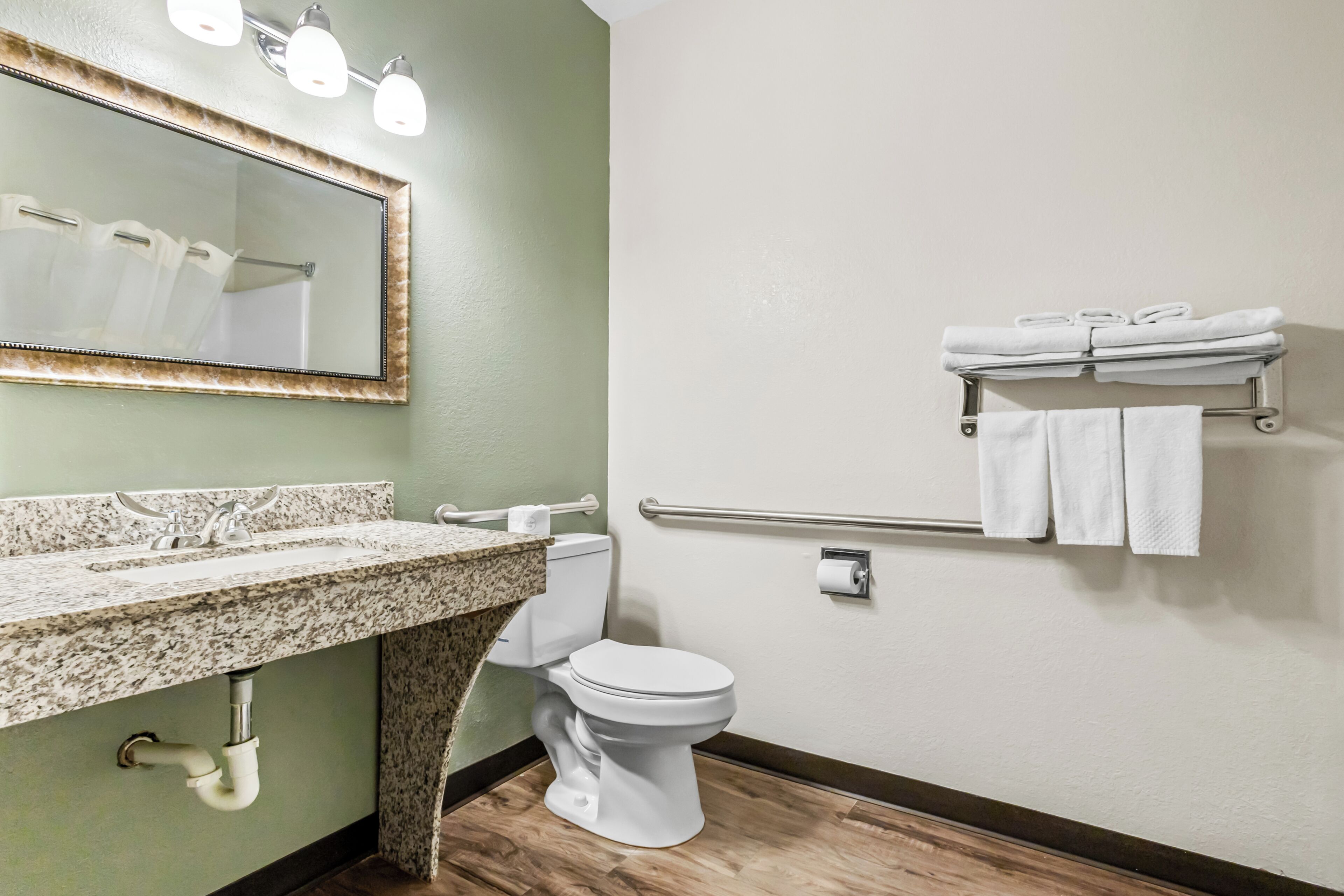 A bathroom with a granite countertop sink, a white toilet, a mirror with lights, and a towel rack with towels.