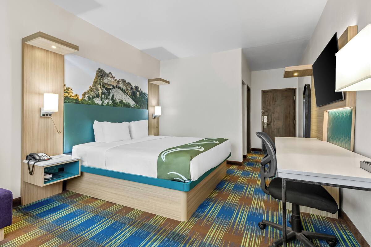A hotel room with a bed, desk, and a painting of a mountain.