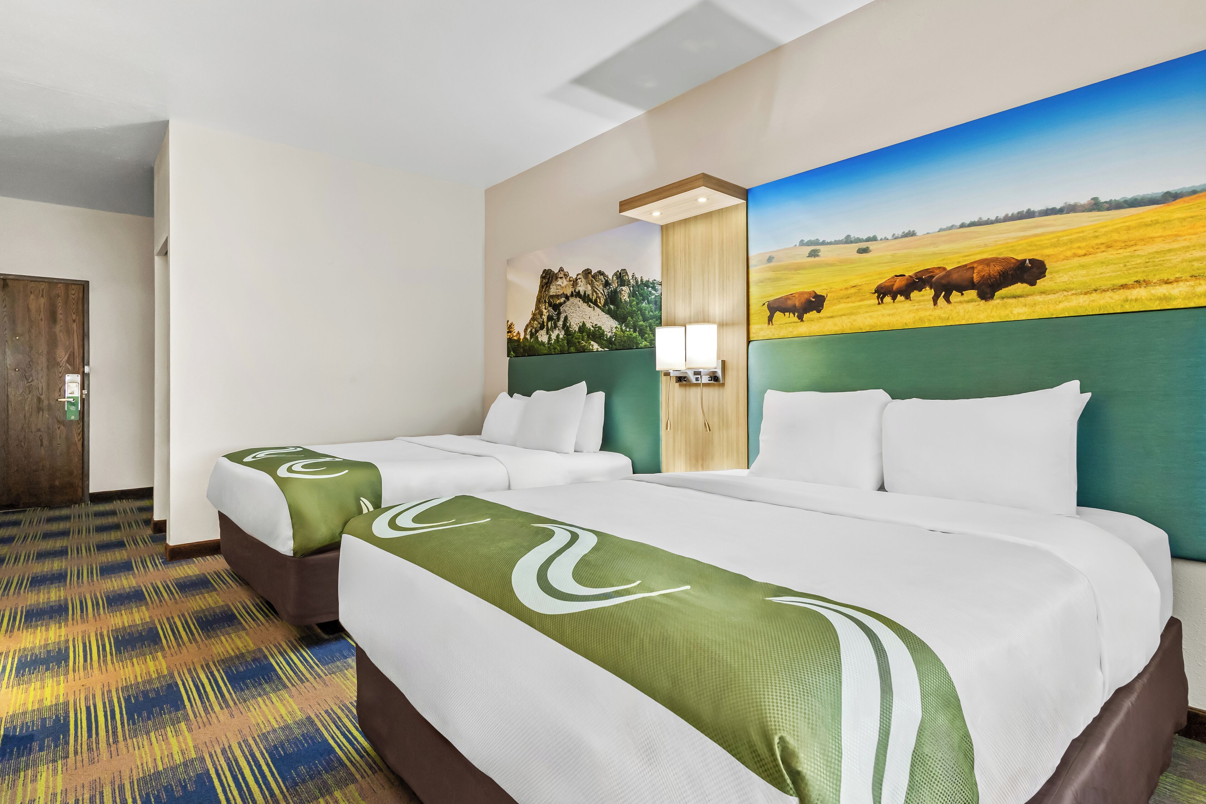 A hotel room with two beds, a large painting of a field with bison, and a wooden door.