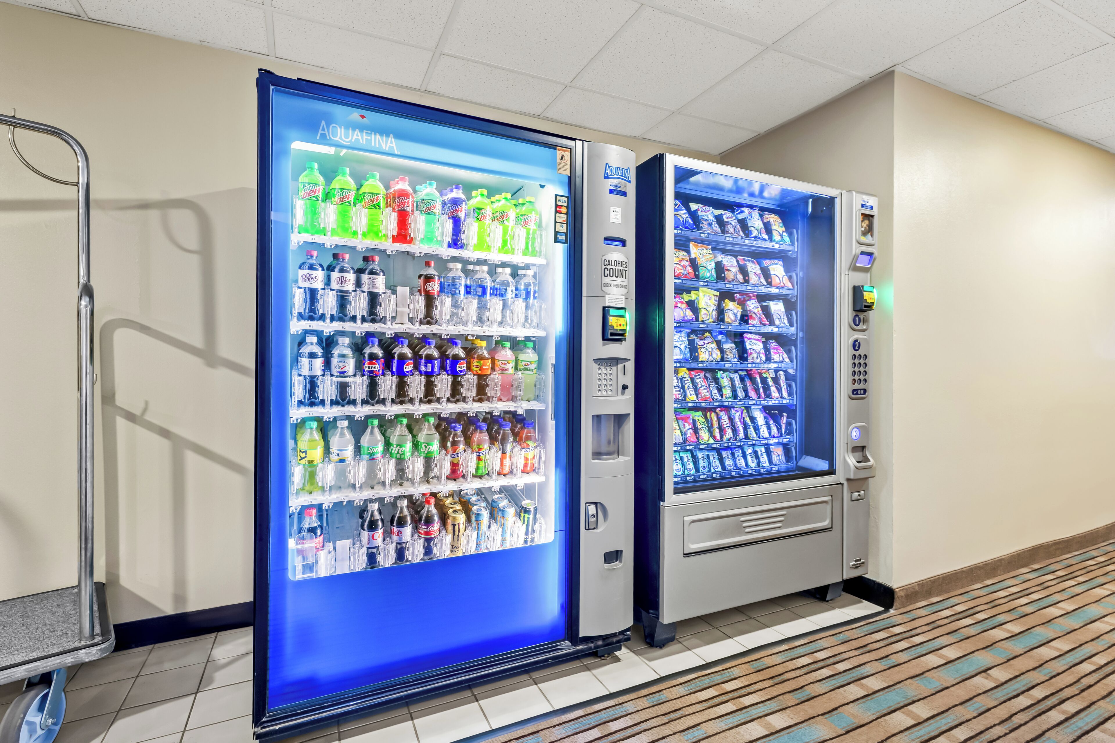 Two self-service beverage vending machines with a variety of drinks and snacks, located in a hallway with a patterned floor.