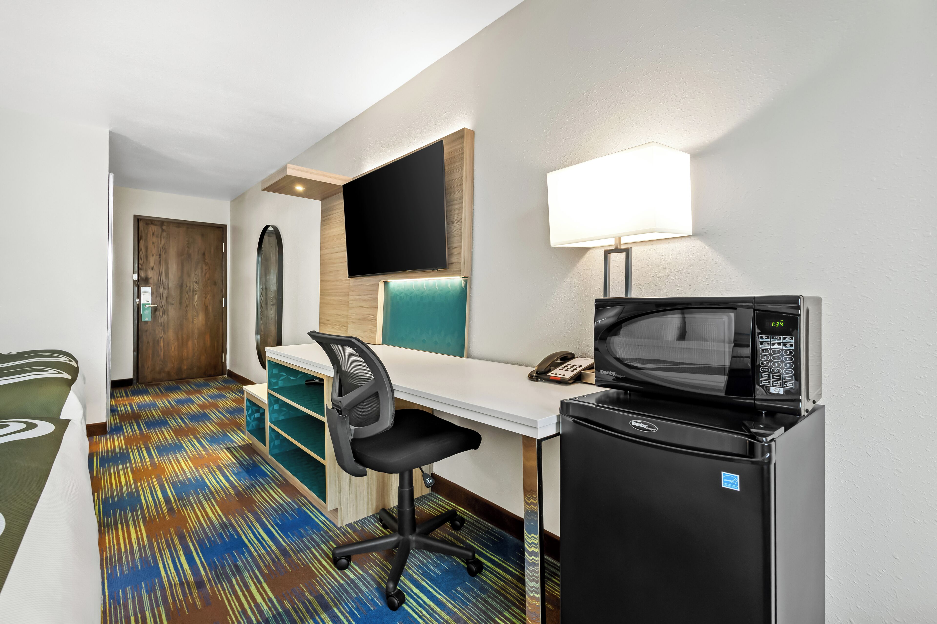 A hotel room with a desk, chair, flat-screen TV, microwave, and a refrigerator.