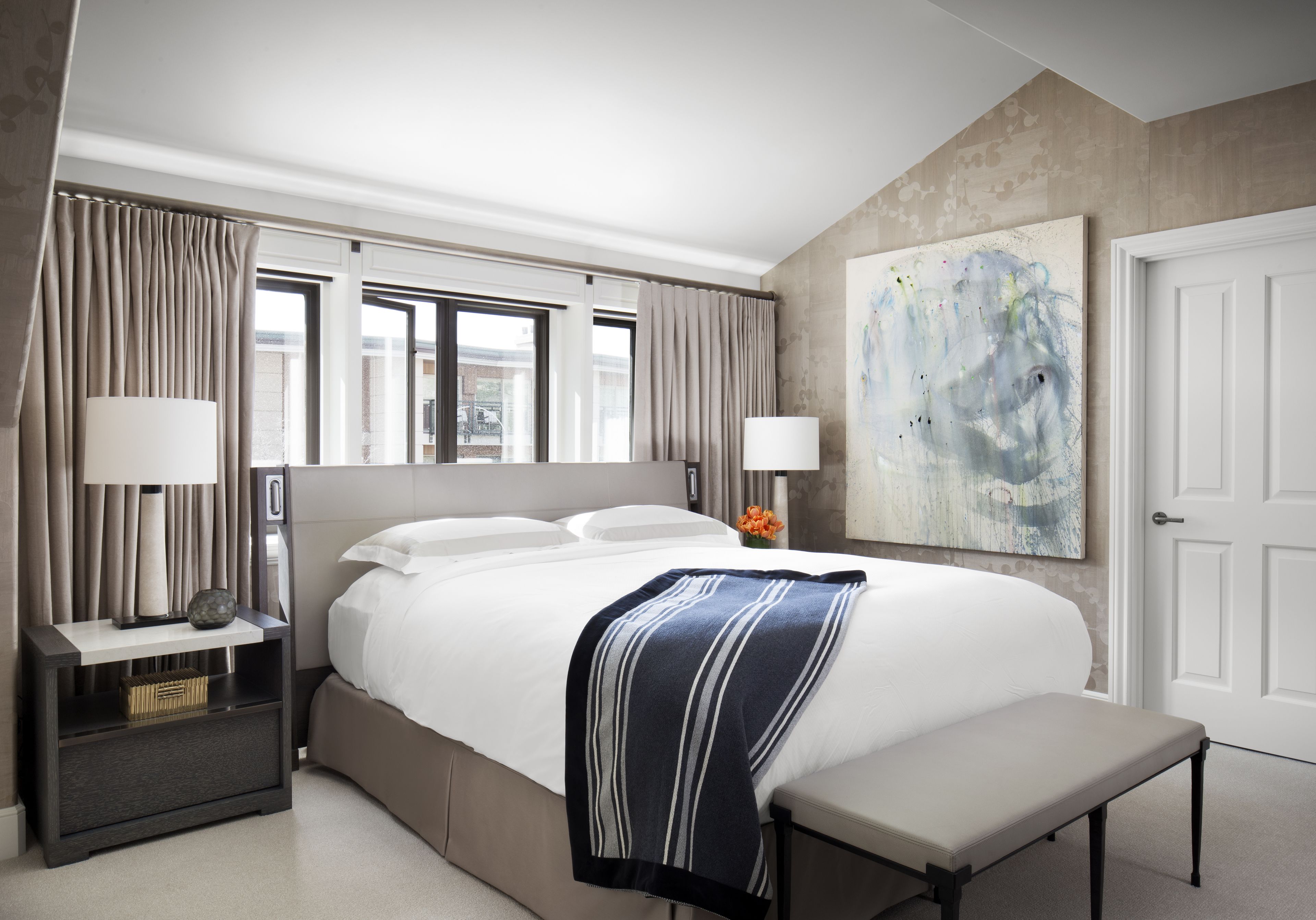 A modern hotel room with a large bed, bedside lamps, a bench, and a painting on the wall.