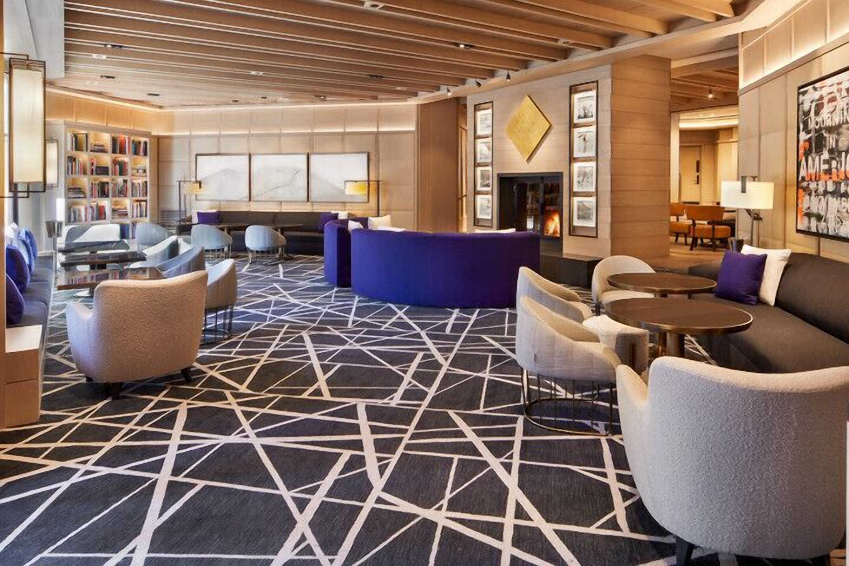 A modern hotel lobby with a geometric patterned carpet, seating areas, a fireplace, and a bookshelf.
