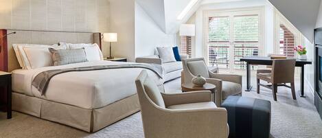 Quarto de hotel moderno com uma cama grande, área de estar com uma poltrona e apoio para os pés, uma mesinha e uma televisão.
