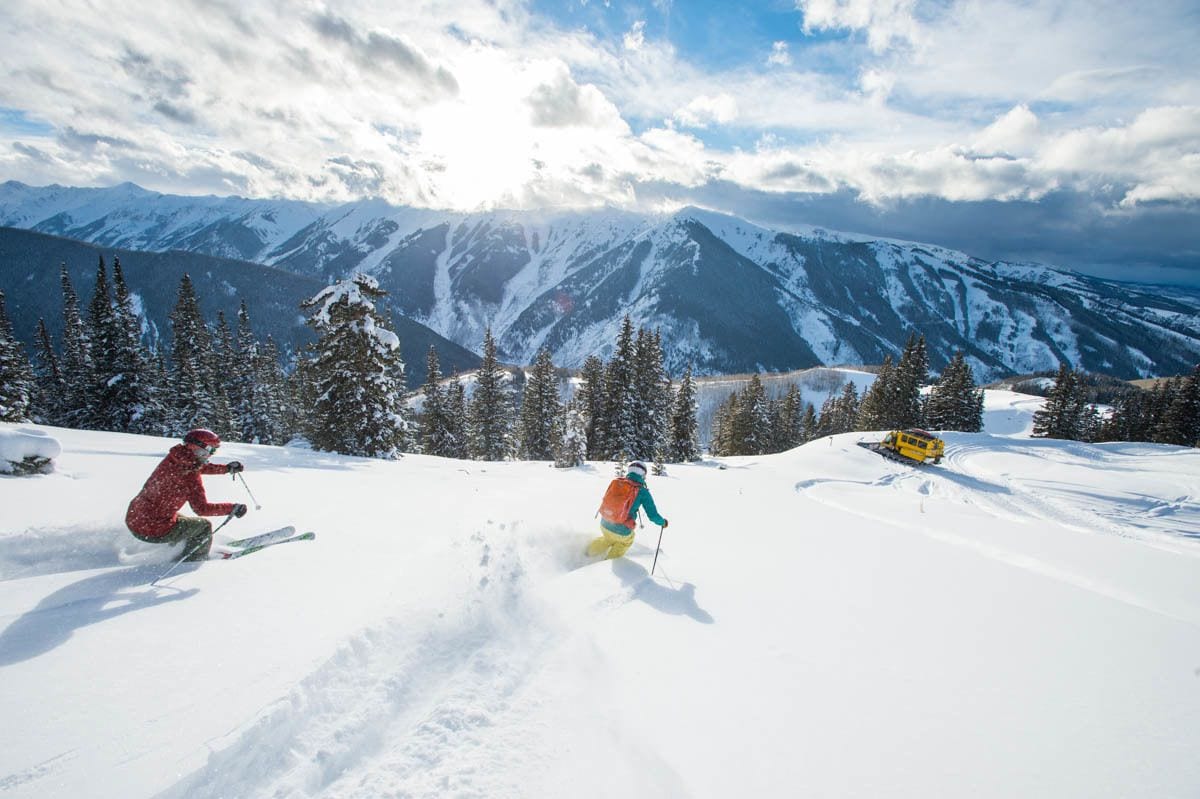 Eine verschneite Berglandschaft mit Skifahrern und einem gelben Fahrzeug.