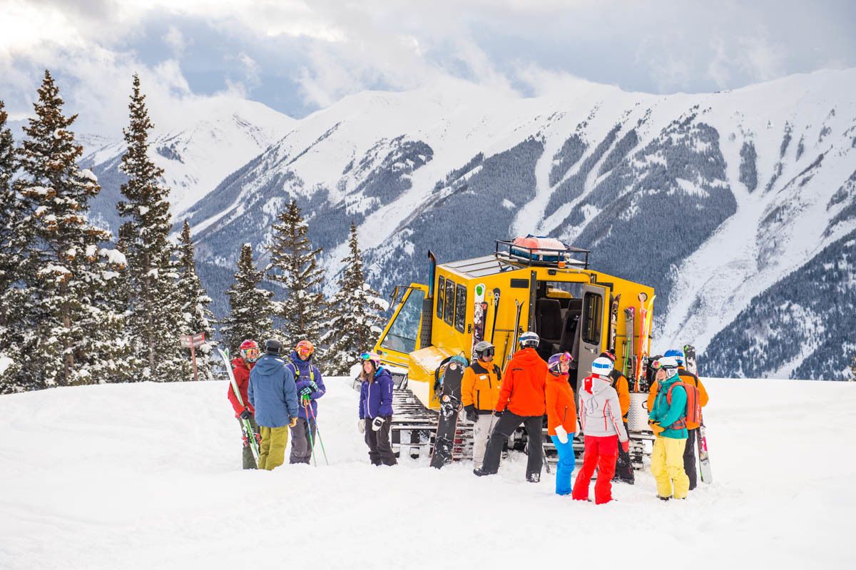 A group of skiers gathered around a yellow vehicle in a snowy mountainous area.