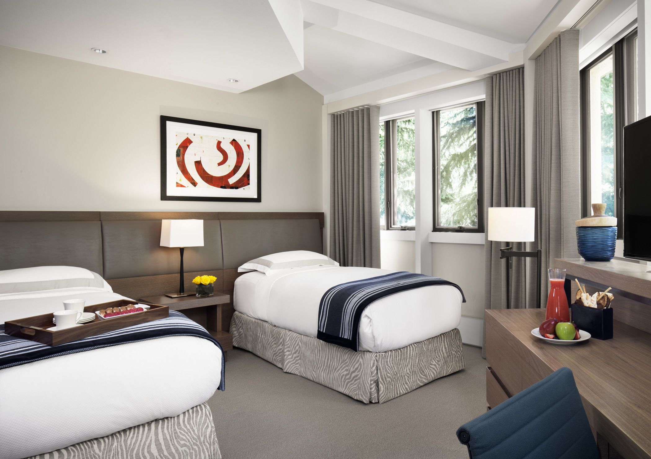 A hotel room with two beds, a desk with a computer, a window with curtains, and a framed artwork on the wall.