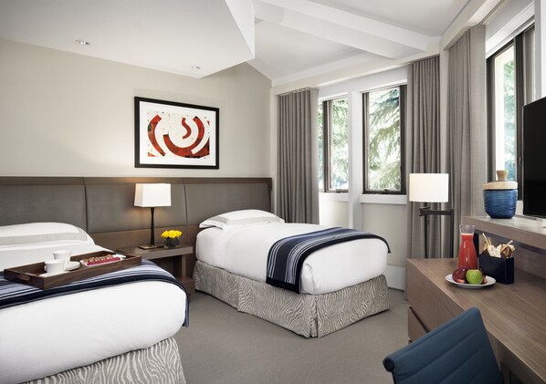 A hotel room with two beds, a desk with a computer, a window with curtains, and a framed artwork on the wall.