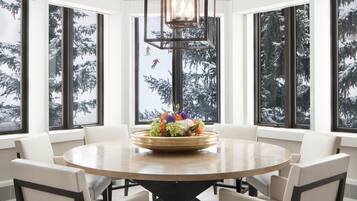 A dining area with a round table, six chairs, a chandelier, and large windows with a snowy view.