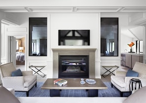 A modern living room with a fireplace, two armchairs, a coffee table with books, and a large TV.