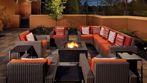 An outdoor seating area with wicker furniture, a central fire pit, and a view of the surrounding buildings. - Courtyard by Marriott State College (State College)