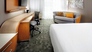 A hotel room with a bed, a desk with a chair, a sofa with cushions, a flat-screen TV mounted on the wall, and a large window with curtains. - Courtyard by Marriott State College (State College)