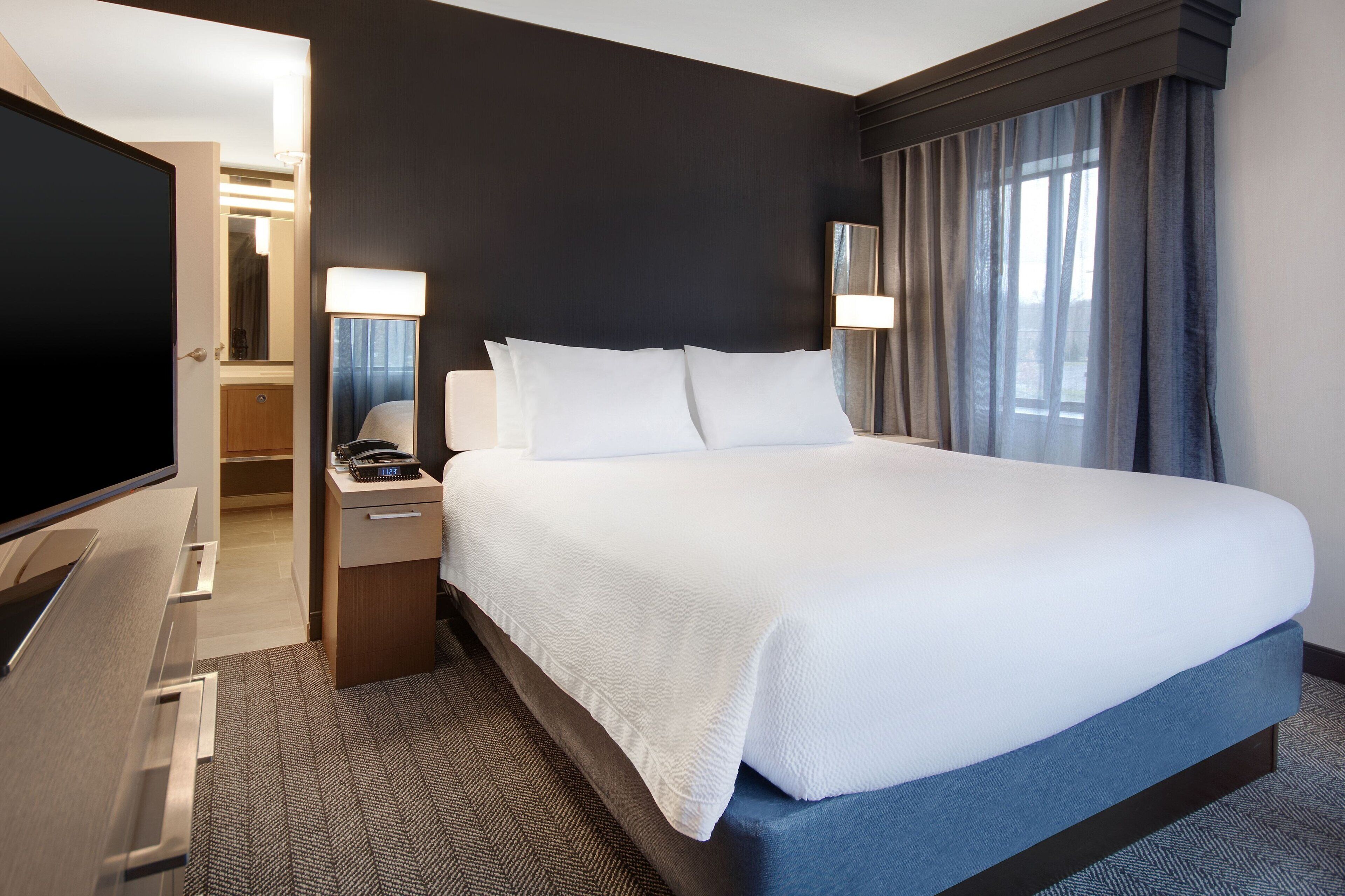 A hotel room with a large bed, a television, a desk with a lamp, and a bathroom visible through an open door.