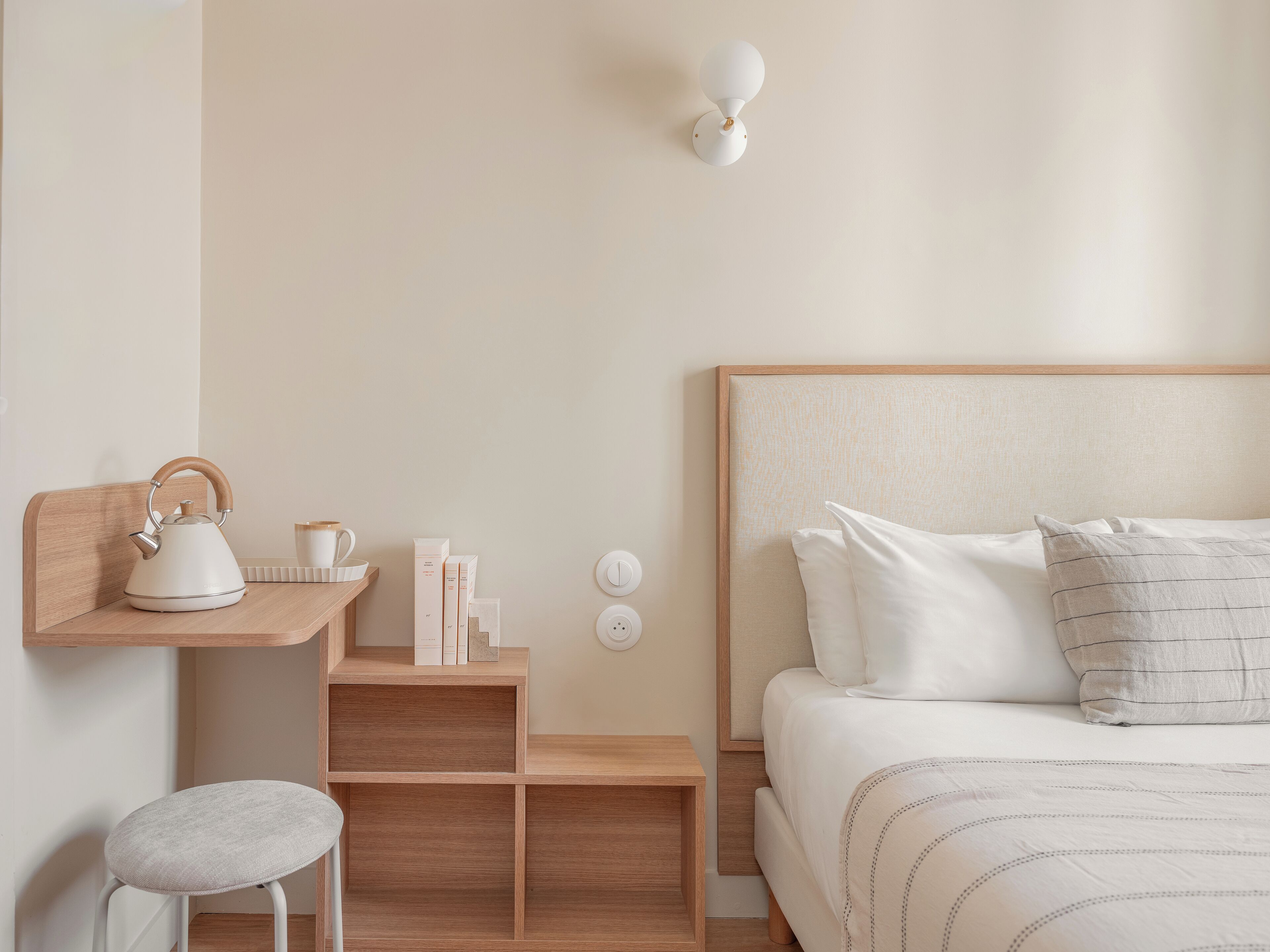 A bedroom with a wooden headboard, a bedside table with a kettle, books, and a lamp.
