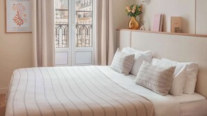 A hotel room with a large bed, a view of the city through a window, and a shelf with books and a vase. - Hôtel Basss (Paris)