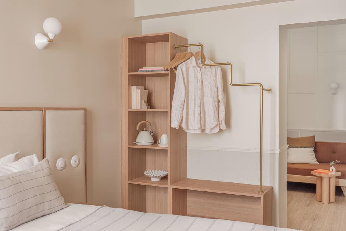 Une chambre moderne avec une tête de lit en bois, une table de chevet avec une bouilloire, une étagère avec des livres et un porte-manteau suspendu, et une vue sur une pièce adjacente.