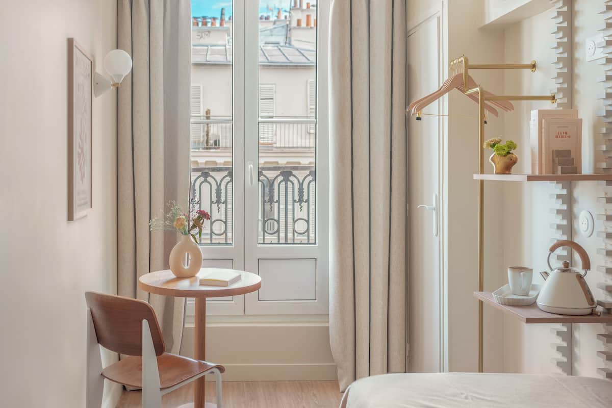 Une chambre d’hôtel avec un lit, une table ronde et une chaise, un vase avec des fleurs, une bouilloire et une étagère avec des livres et une plante.