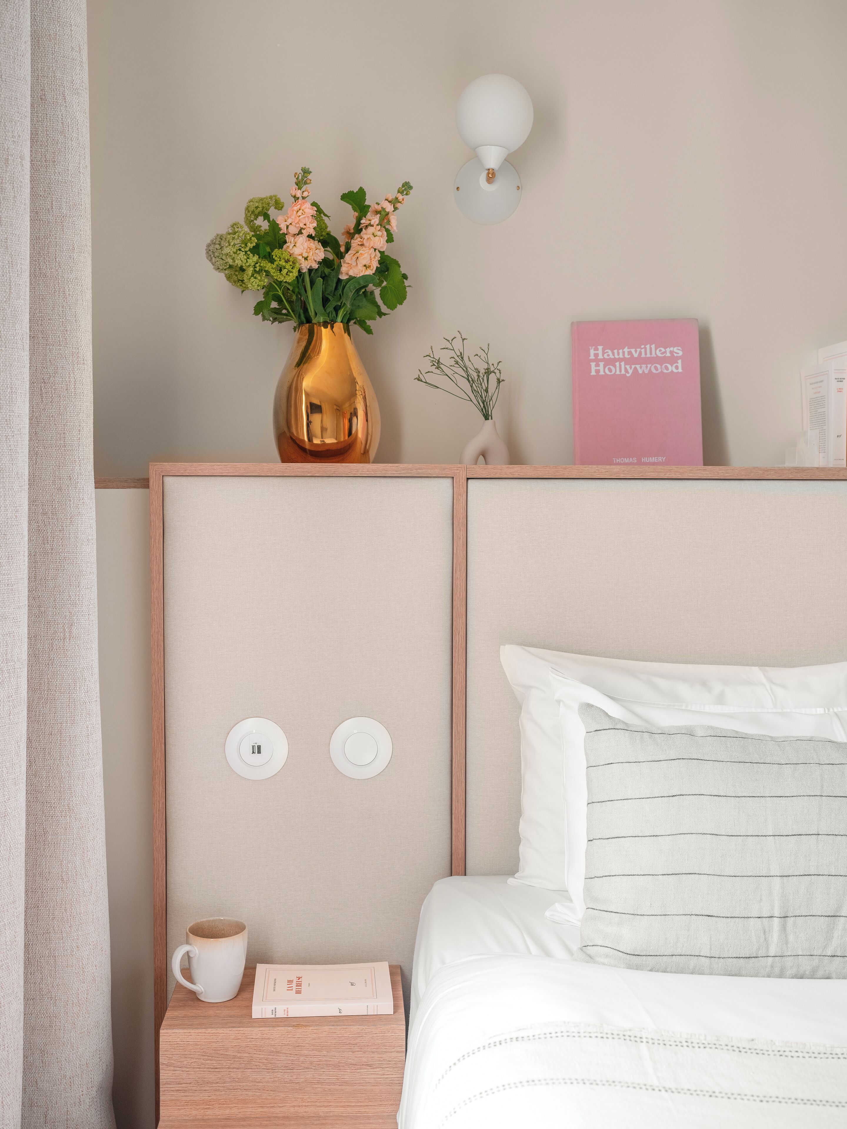Un dormitorio ordenado con una cama, una mesita de noche y una lámpara de pared. En la mesita de noche hay un libro titulado 