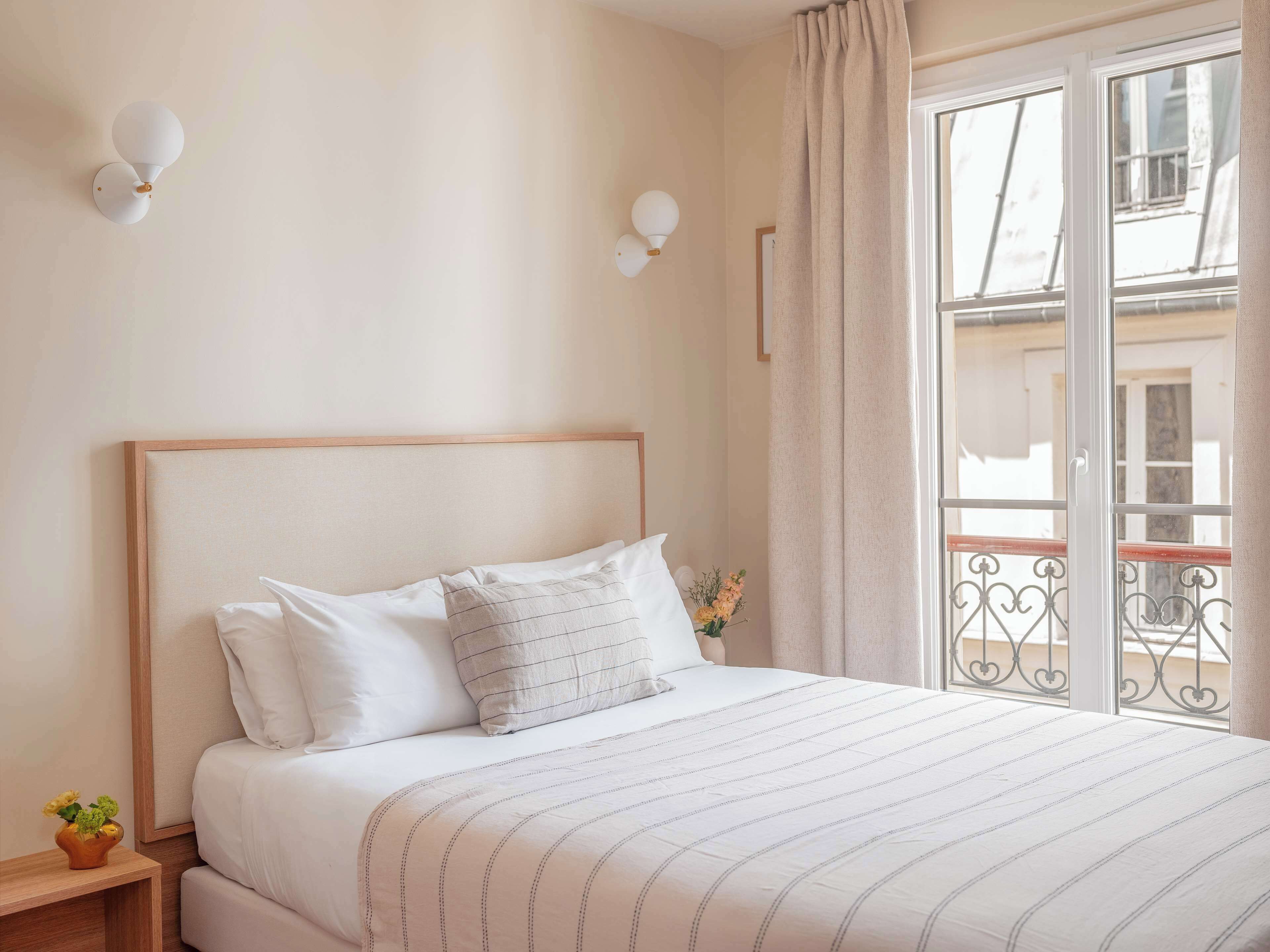 A bedroom with a bed, bedside table, wall-mounted light fixtures, and a window with a view of buildings.